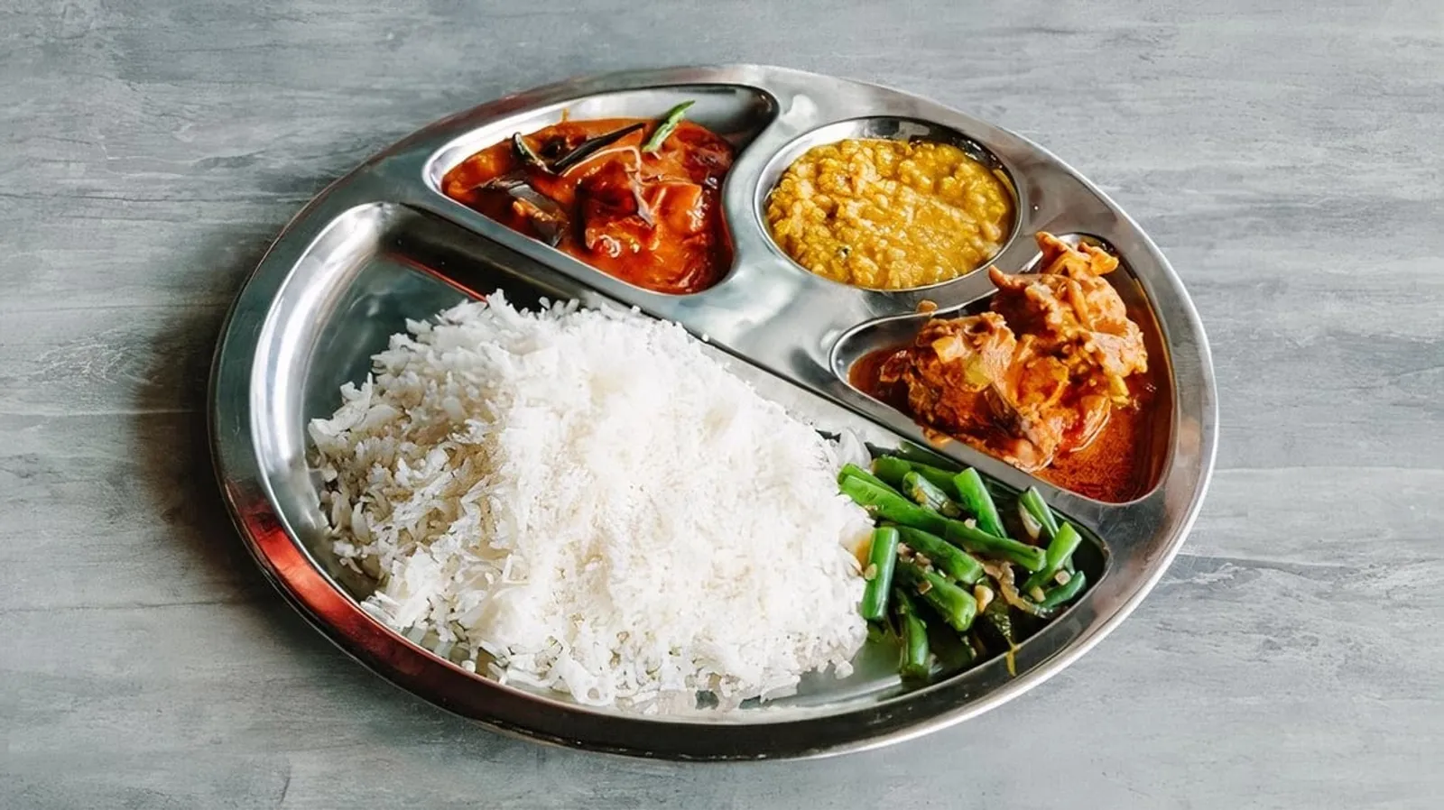 South Indian steel thali plate with rice, two curries, dhal and stir-fried green beans on grey stone