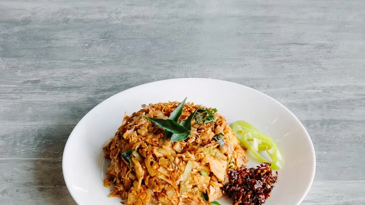 Editorial shot of kothu fried rice on white plate with pandan leaf, cucumber and chilli sambal on grey stone