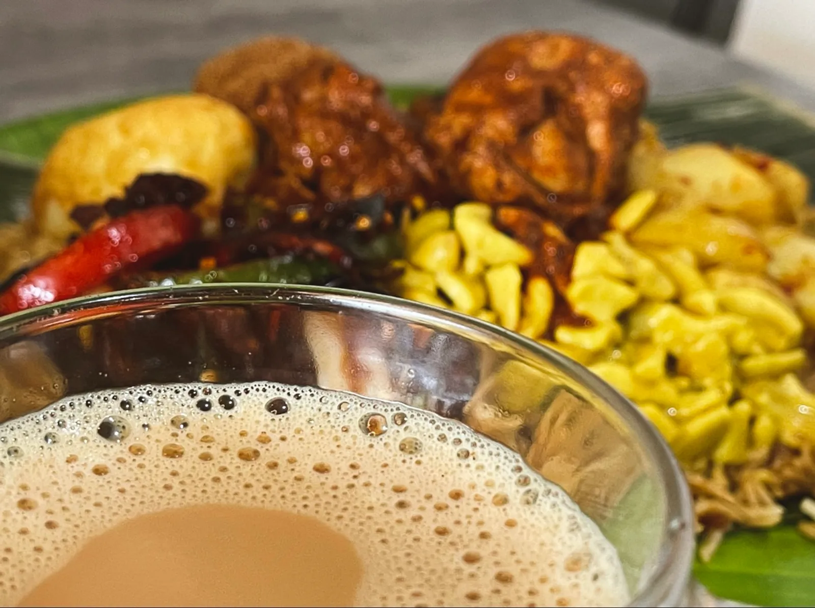 Teh tarik in glass bowl with biryani and vadai fritters blurred behind on banana leaf