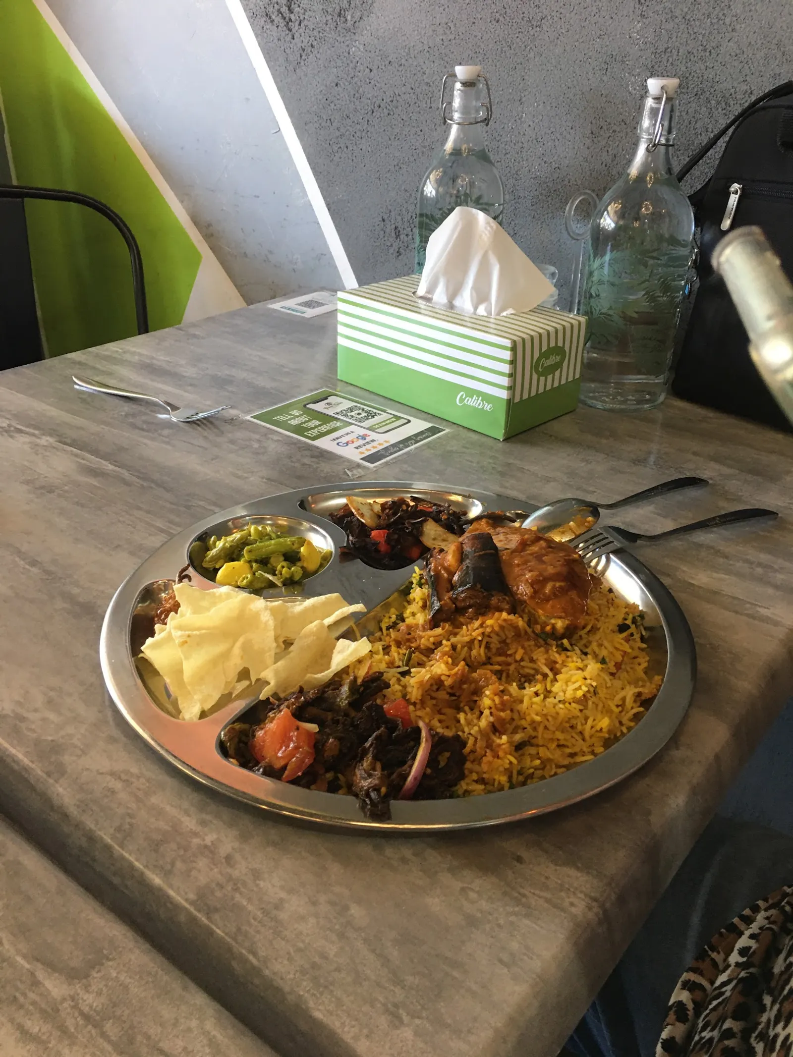 Stainless steel thali with biryani, chicken and curry on concrete outdoor table with water bottles